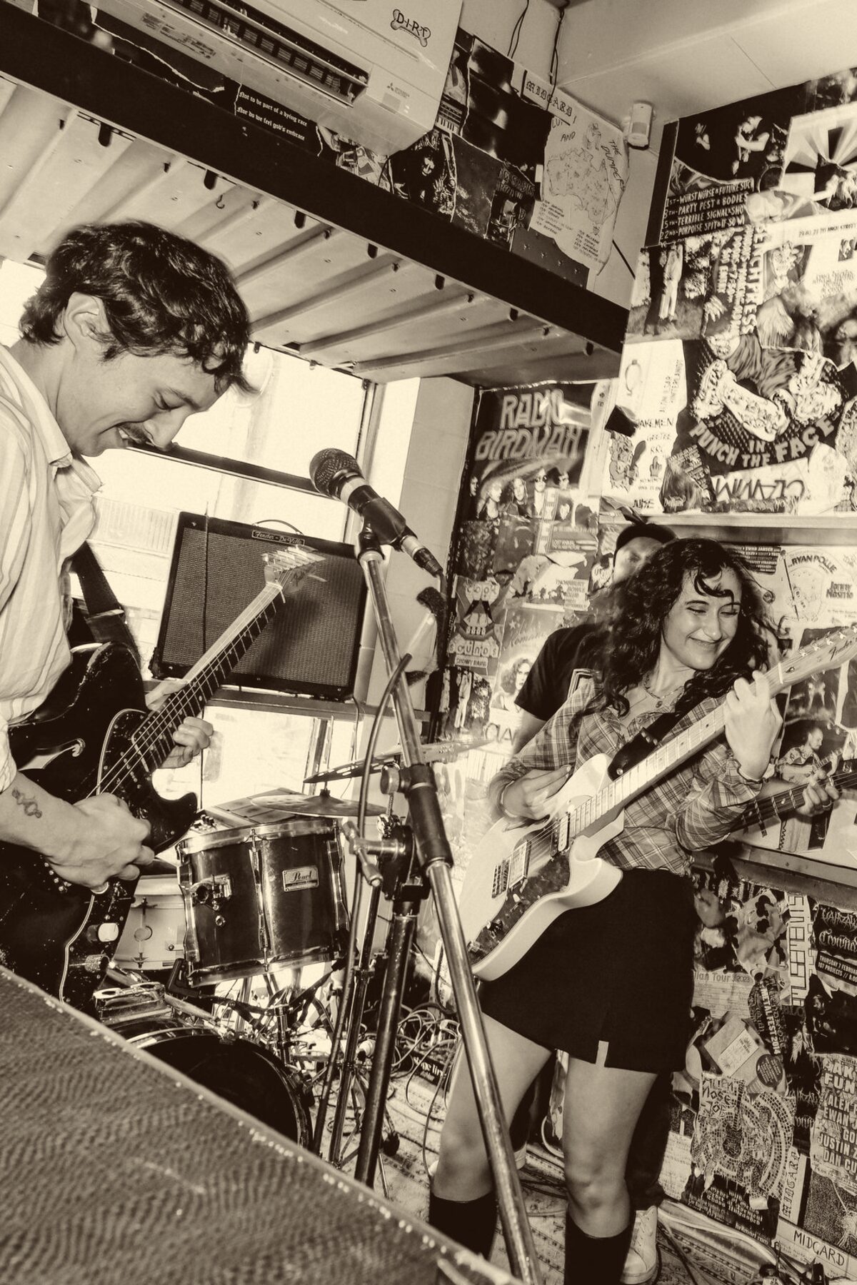 A guitarist grinning mid-song in a poster-covered room, a bandmate beside them